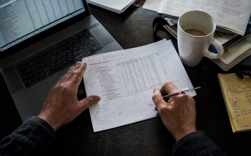Planilha impressa anotada à mão ao lado de um laptop com uma planilha aberta